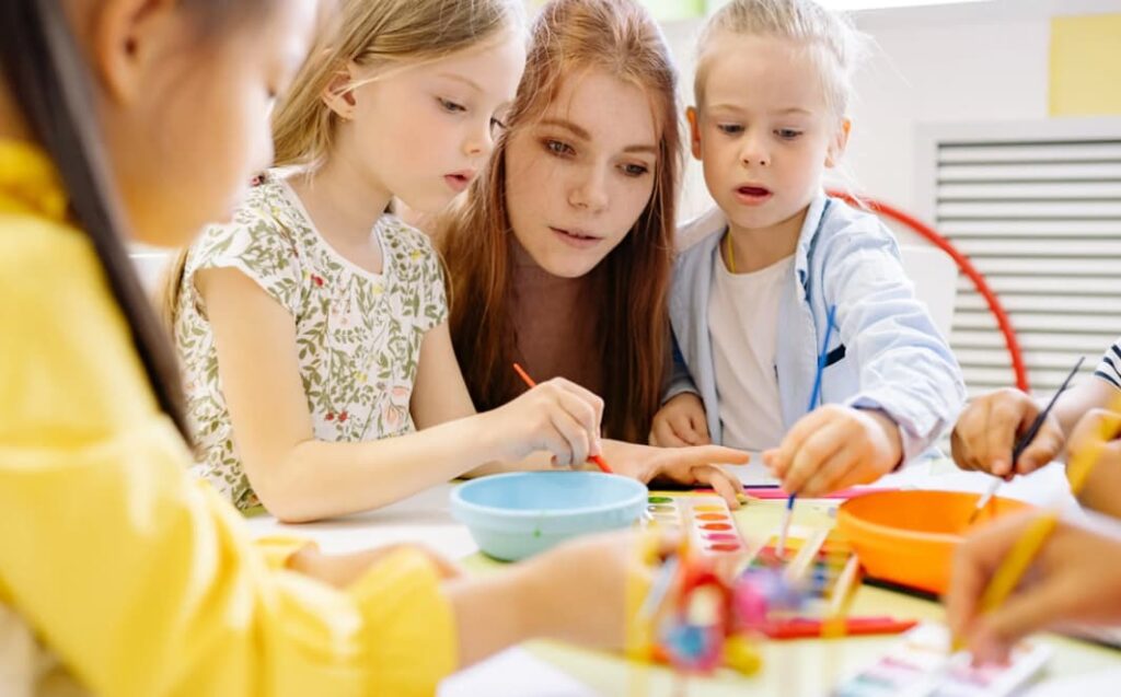 A young teacher with red hair is sitting at a table with preschool children, helping them with crafts