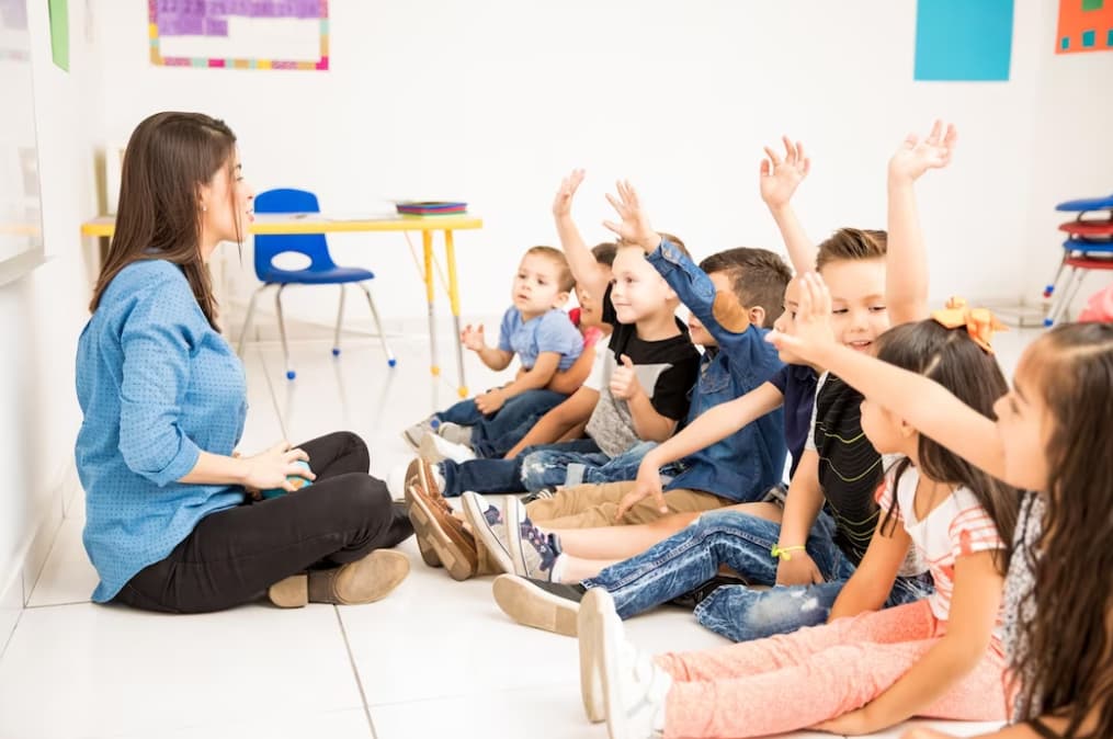 woman in a blue shirt sits in front of the children with their hands up