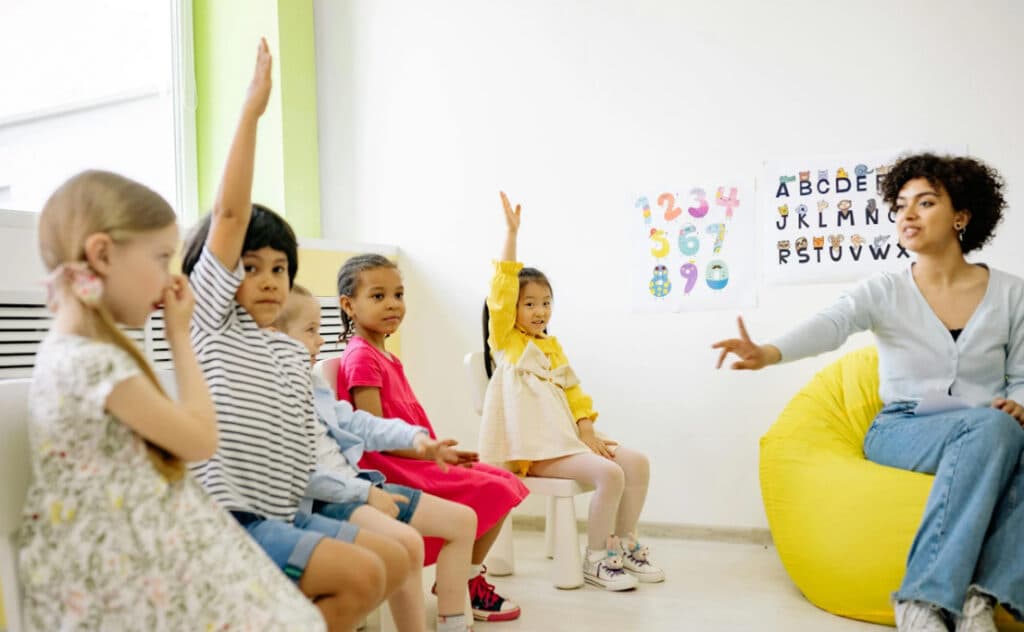 A diverse group of children with raised hands in a classroom, with a teacher pointing to one of them