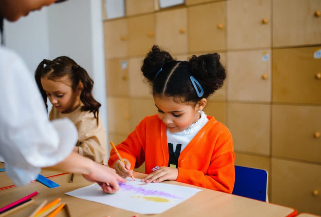 A young girl with puffy hair drawing, with another child and teacher nearby