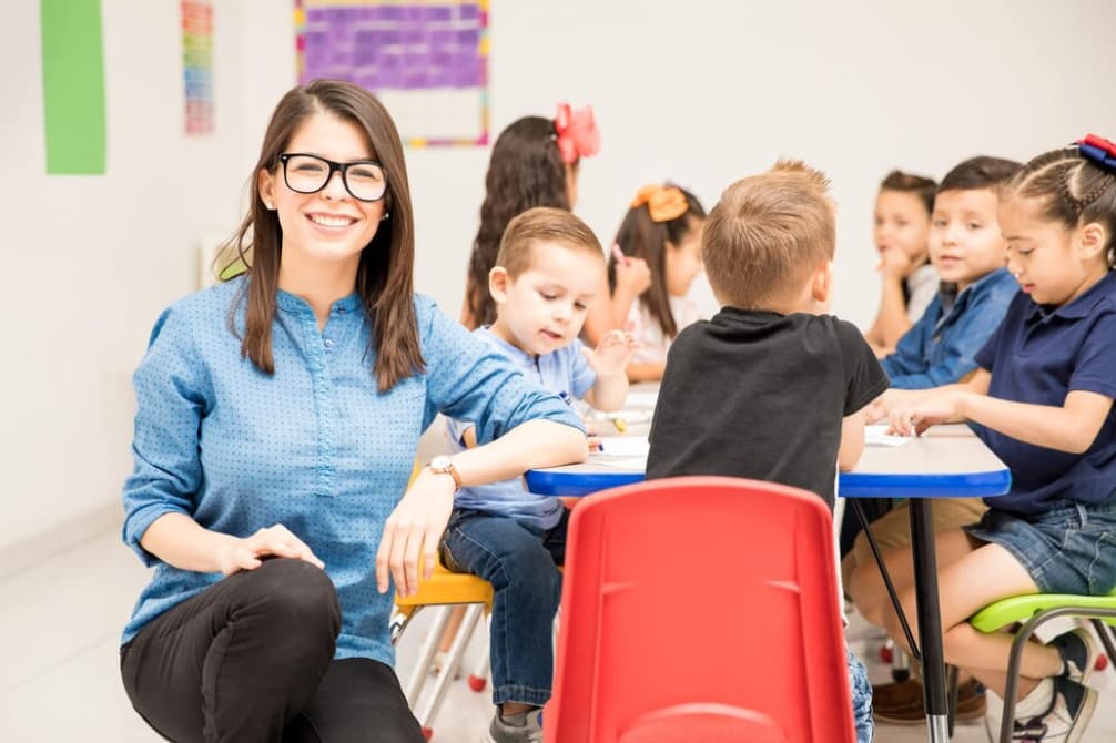 A smiling teacher sits with young students at tables in a bright classroom